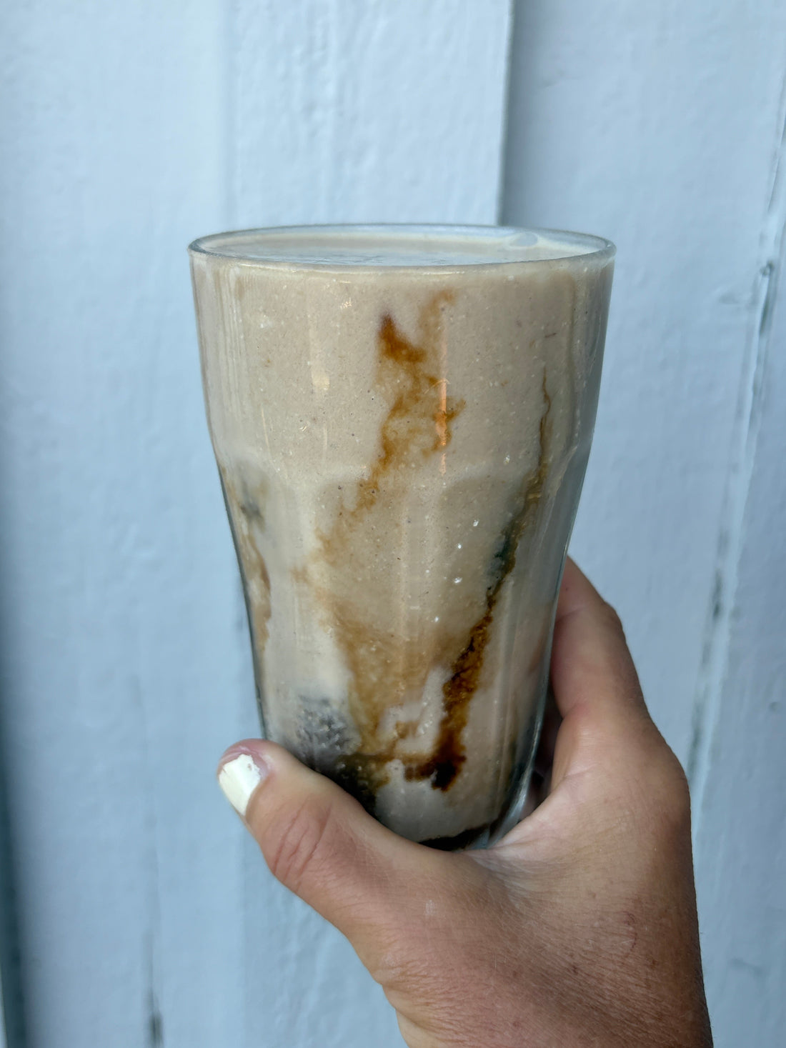 Hand som håller ett högt glas proteinshake med kaffe, jordnötssmör och banan, gjord på protein från POW