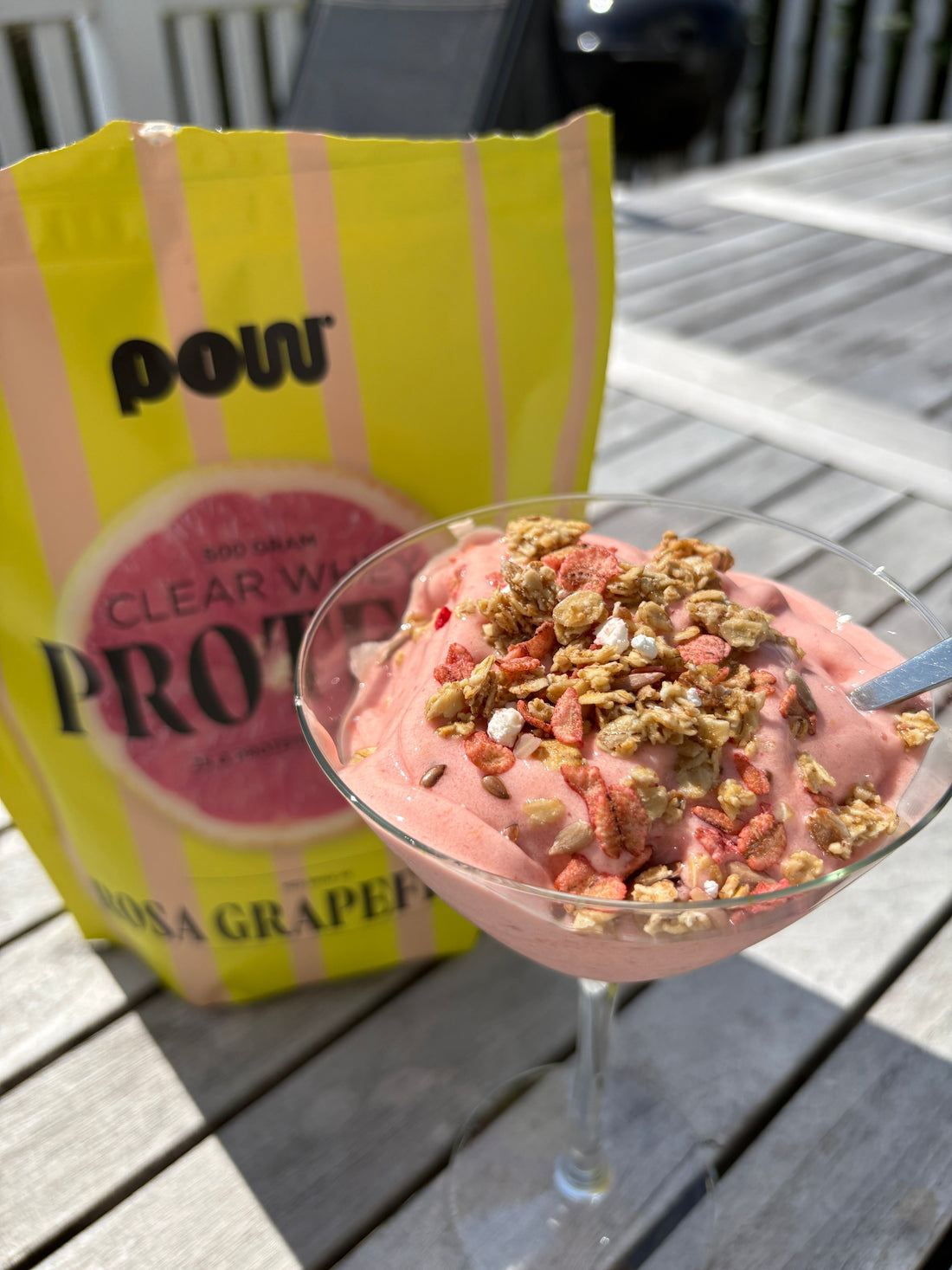 Rosa himmel-proteinsmoothie i glas toppad med granola, bredvid en påse POW Clear Whey Protein