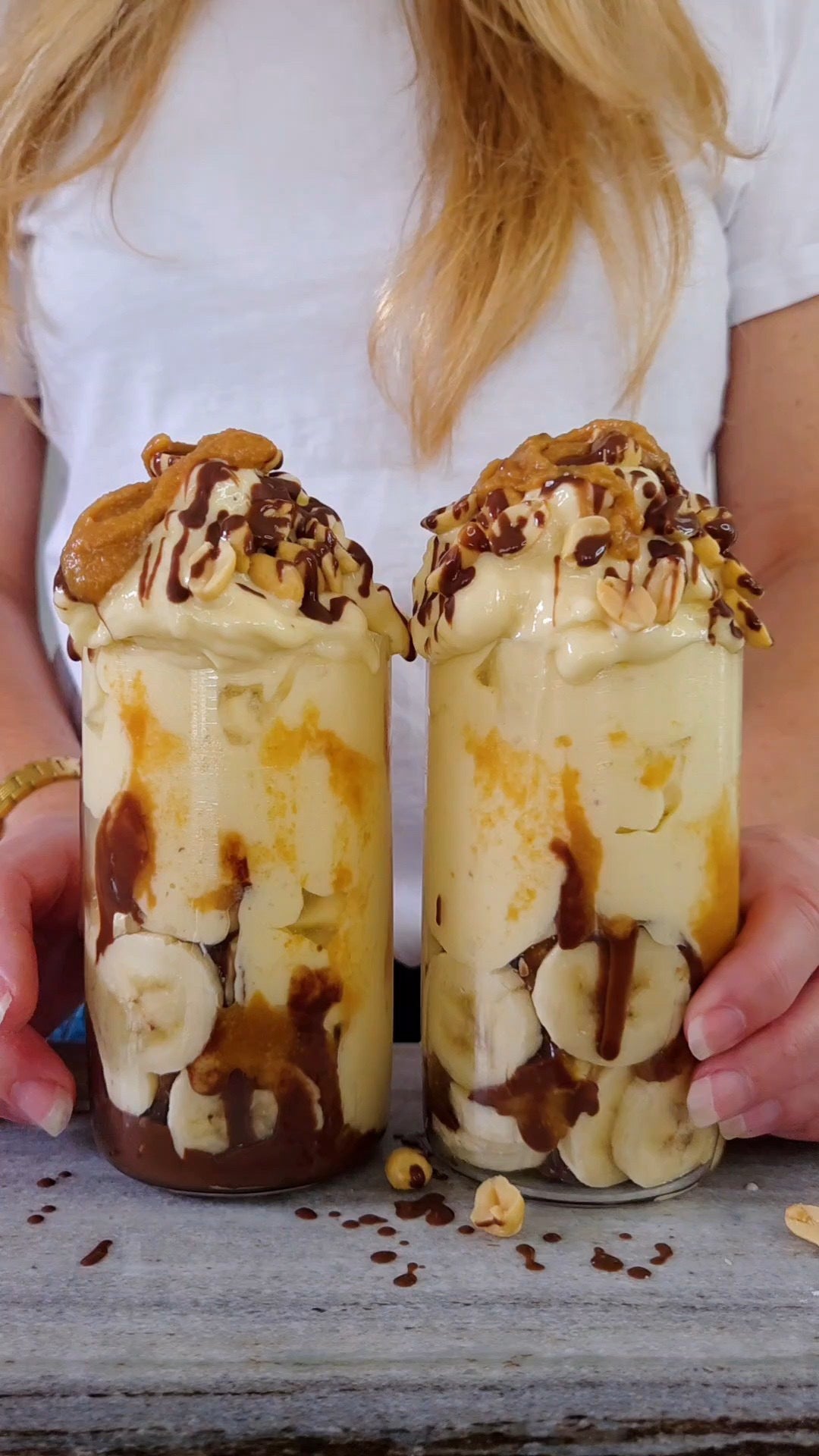 Två höga glas med Snickers-nicecream på kaseinprotein vanilj, varvad med banan, chokladsås och kolasås, toppad med jordnötter