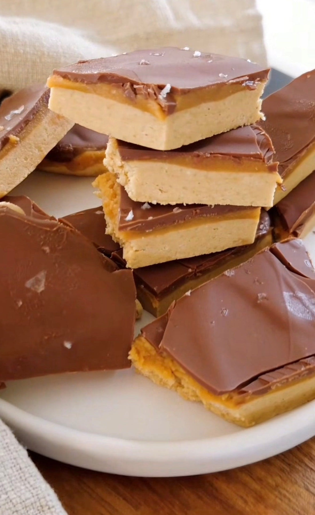Staplade bitar av no-bake proteinbark med lager av ljus proteinbas, kola och mörk chokladöverdrag, strödda med flingsalt på en vit tallrik.