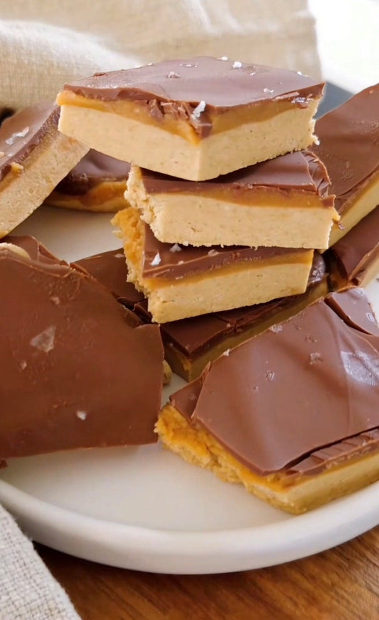 Staplade bitar av no-bake proteinbark med lager av ljus proteinbas, kola och mörk chokladöverdrag, strödda med flingsalt på en vit tallrik.