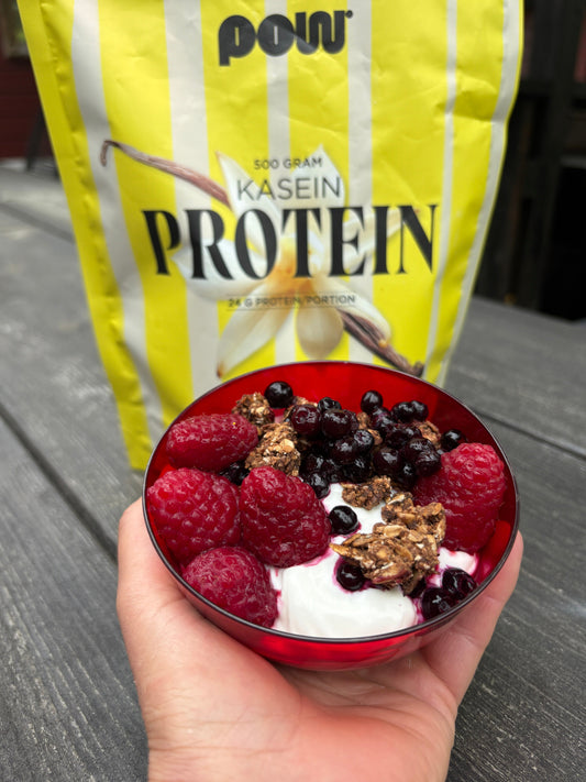 Röd skål med krämig proteinrik yoghurt toppad med hallon, blåbär och granola, hållen av en hand utomhus med en gul POW Kasein Protein-påse i bakgrunden